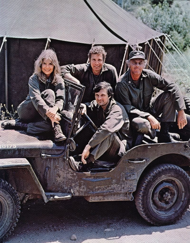 American actor, director and writer Alan Alda in the driving seat of a jeep, surrounded by Loretta Swit and other cast members of the hit television show M.A.S.H, in costume as members of a US Army medical corp. 