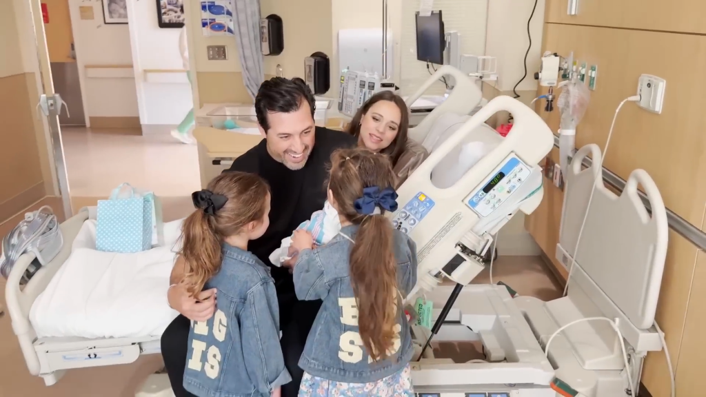 With a fully made-up Jinger Duggar peeping over his shoulder, Jeremy Vuolo holds baby Finnegan while Felicity and Evangeline look on at their baby brother at the end of March 2025.