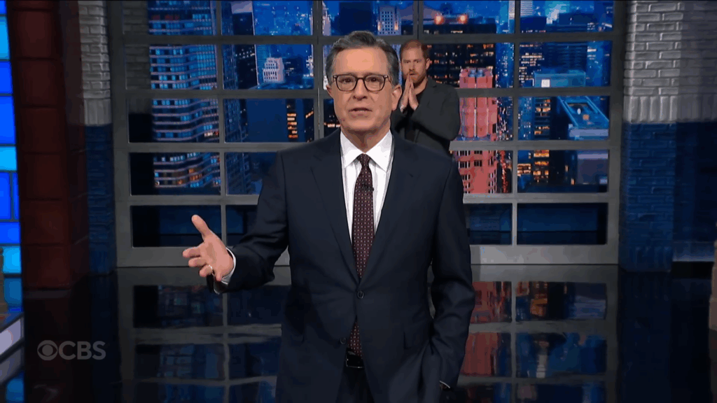 Stephen Colbert addresses the audience with Prince Harry behind him.